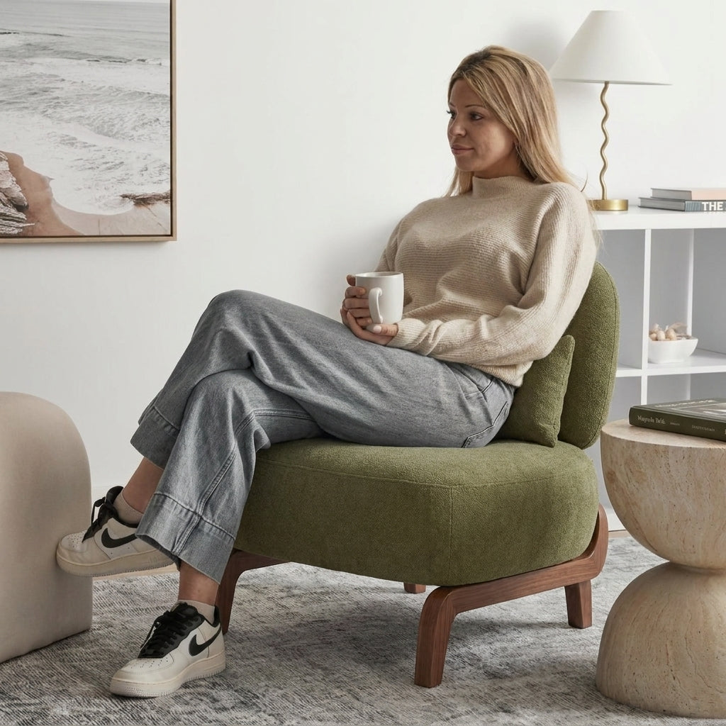 A woman sits in a green upholstered armchair with wooden legs, holding a white mug in a bright, modern living room. She is wearing a beige sweater, gray wide-leg jeans, and black-and-white sneakers.