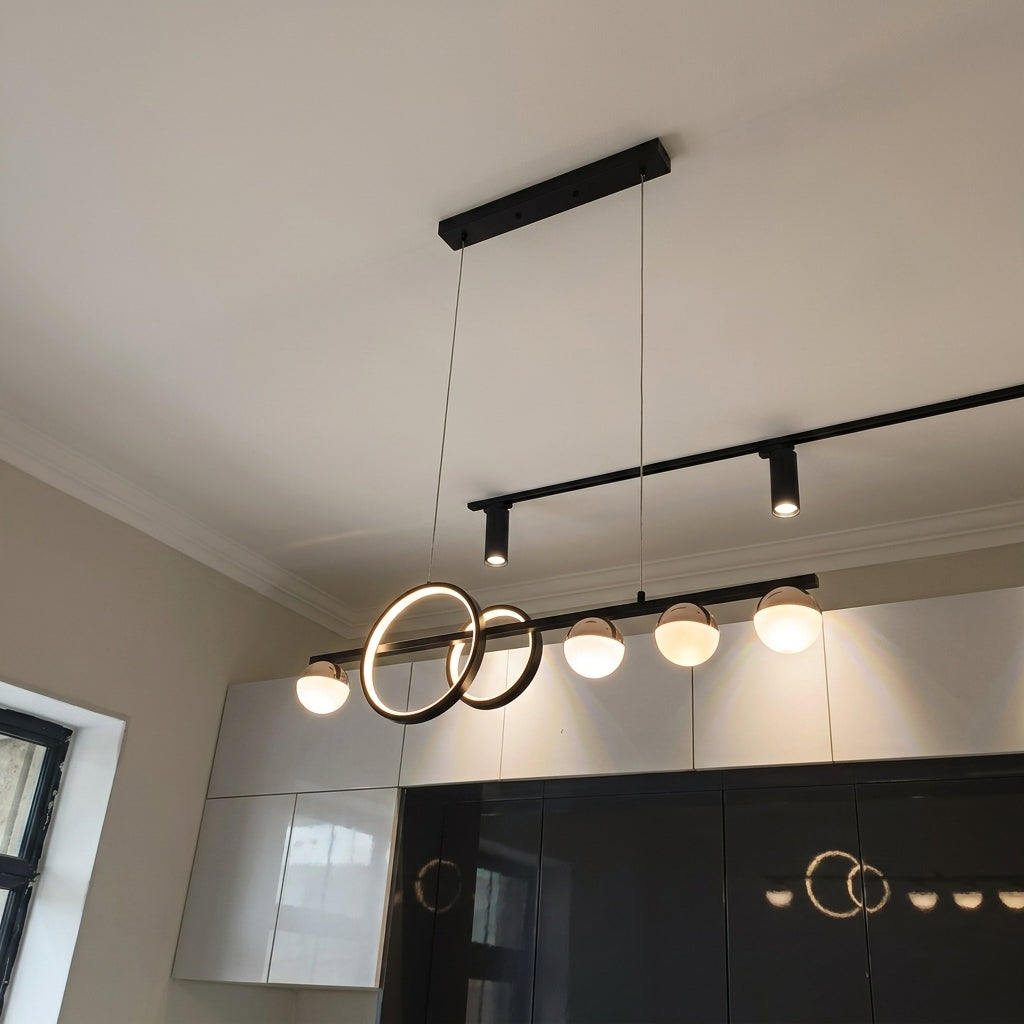 Modern black metal linear pendant light fixture with two interlocking circular rings and five glowing globe bulbs, shown hanging in a contemporary kitchen above white and black cabinets.