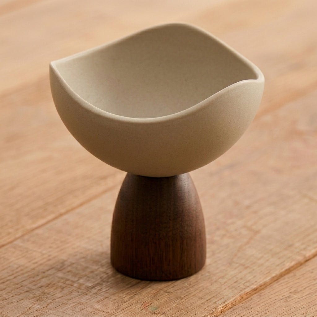A ceramic and wood pedestal bowl with a rounded, slightly wavy rim, set on a light brown wooden surface.