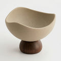 A modern ceramic bowl with a wavy, organic shape in a light beige speckled glaze, resting on a round dark brown wooden pedestal base against a clean white background.