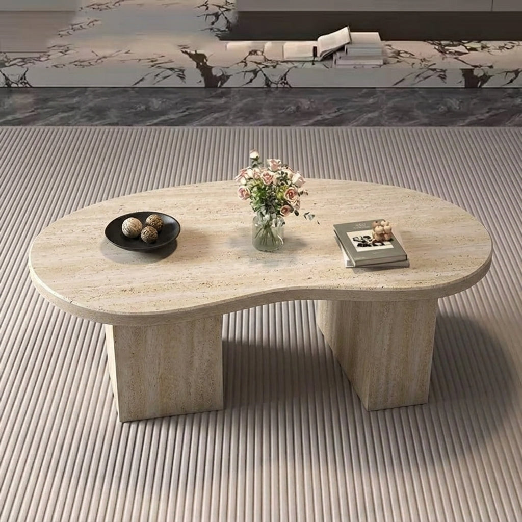An organic-shaped beige travertine stone coffee table with a textured pedestal base, styled with a black bowl, a vase of flowers, and a book on a ribbed light beige rug.