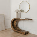 A rustic crescent-shaped wooden console table in a warm medium brown finish, styled with a beige table lamp and books, set against a white paneled wall with a gold-framed mirror.