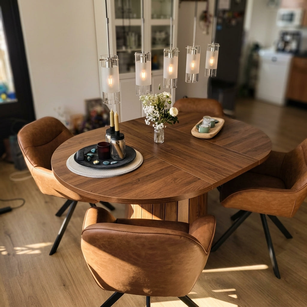 A round wooden dining table with a central pedestal base, surrounded by four brown leather swivel chairs with black metal legs. The table is styled with a floral centerpiece, a candle holder, and a small tray, set in a bright, modern dining room with warm sunlight streaming in from the side.