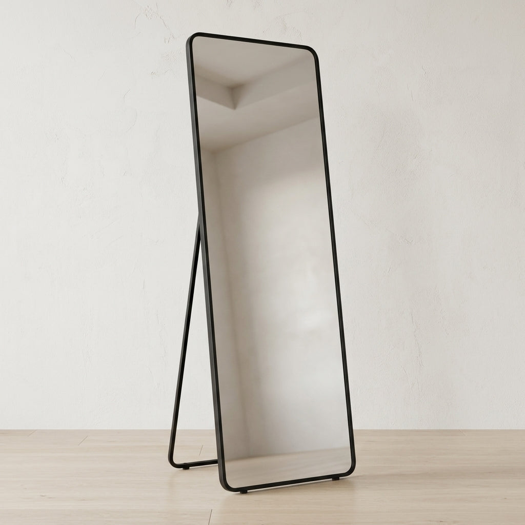 A full-length floor mirror with a slim black metal frame and stand, set against a neutral off-white wall and light wood floor.