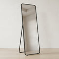 A full-length floor mirror with a slim black metal frame and stand, set against a neutral off-white wall and light wood floor.