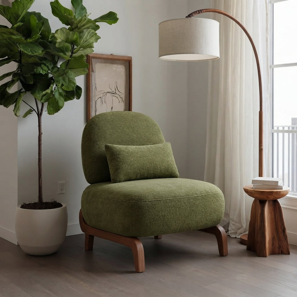An olive green upholstered accent chair with a matching pillow and curved wooden legs, styled in a bright room next to a potted fiddle-leaf fig plant, a floor lamp, and a small wooden side table.