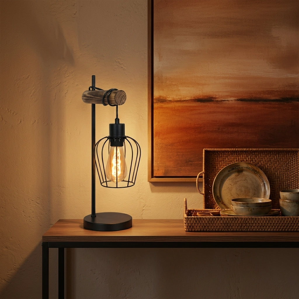 A modern industrial table lamp with a black metal wire cage shade and a single exposed Edison bulb, shown lit on a wooden console table in a warm, styled interior setting.