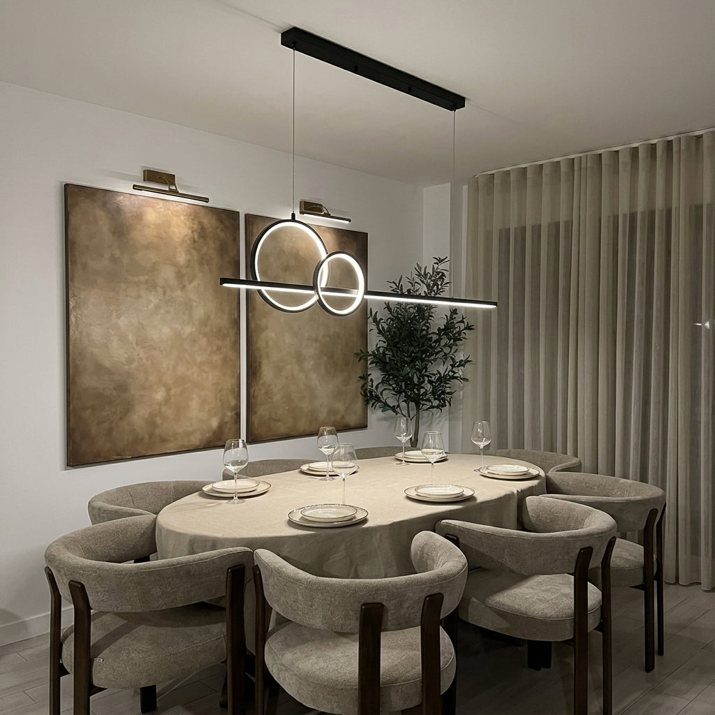 Modern dining room featuring a linear LED pendant light with interlocking circular rings hanging over a round dining table with upholstered chairs. The room has warm neutral walls, textured gold-toned wall art, and sheer curtains.