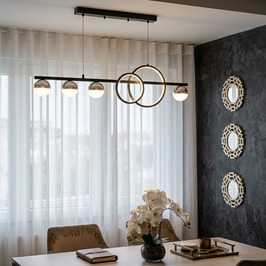 Modern linear pendant light with three spherical glass shades and two interlocking circular metal rings, hanging in a contemporary dining room over a table with chairs and a dark textured wall.