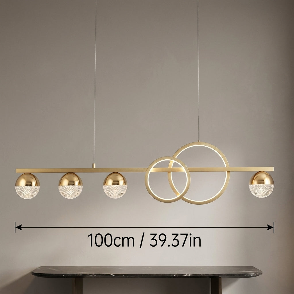 Modern linear pendant light fixture with five gold-toned spherical shades and two interlocking circular gold rings, shown hanging against a gray wall with a dimension marker indicating 100cm (39.37 inches) width.