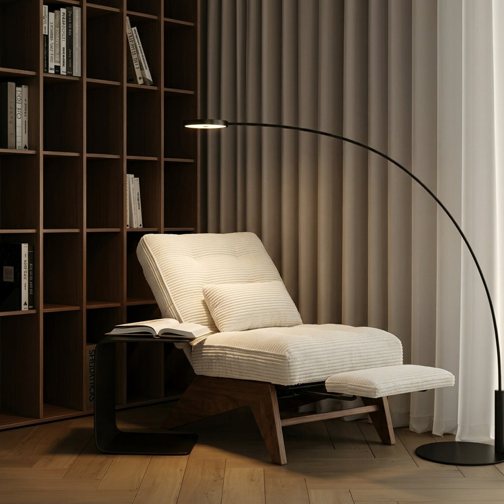 A modern lounge chair with cream ribbed cushions and a wooden base, styled in a cozy room with a black arc floor lamp, a small side table with an open book, and a dark wood bookshelf in the background.