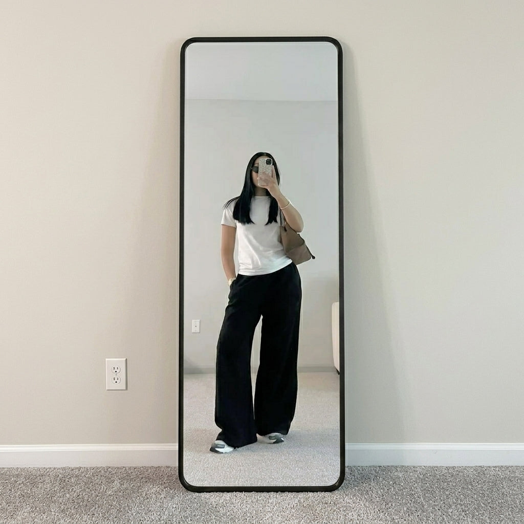 A tall floor mirror with a black frame stands against a beige wall on a gray carpet. The mirror reflects a person taking a mirror selfie wearing a white t-shirt, black wide-leg pants, and white sneakers.