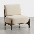 A modern accent chair with textured cream bouclé upholstery and a dark brown wooden frame, shown on a light gray concrete floor against a white studio background.