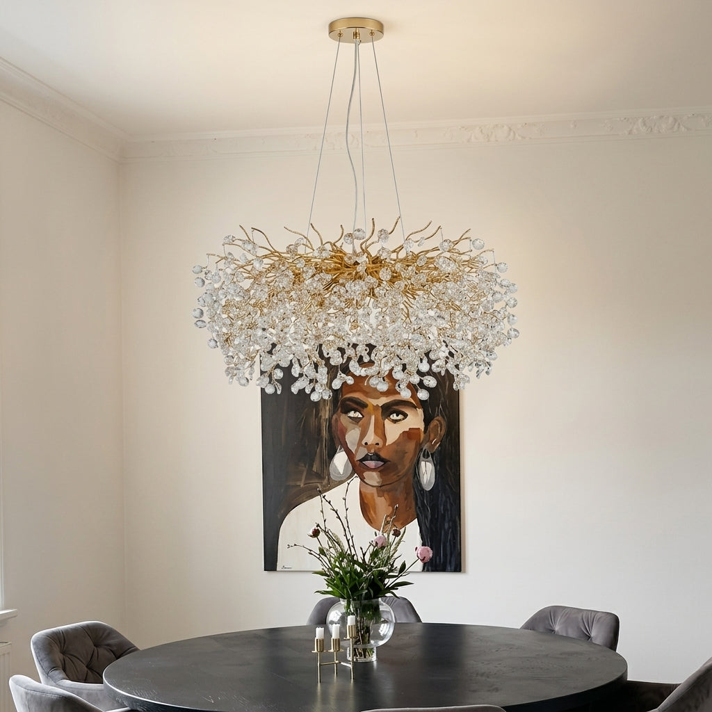 A large, modern chandelier with a gold-toned branching frame and numerous clear crystal droplets, hanging in a dining room above a round black table and in front of a framed portrait on a beige wall.