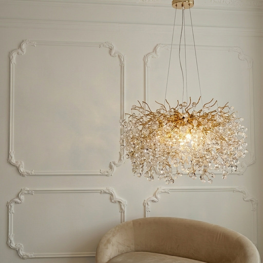 A modern tree-of-life style chandelier with a gold branch structure and clear crystal droplets, illuminated in a luxury room setting with a beige velvet armchair and ornate white wall molding.