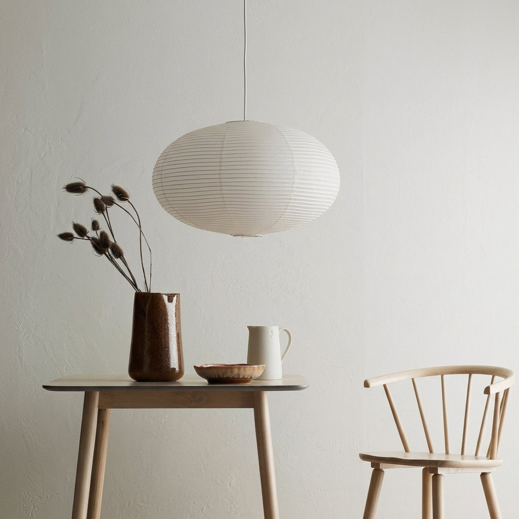 A modern white ribbed paper lantern pendant light hangs over a minimalist wooden table with a ceramic vase, dried stems, and a small bowl in a bright, neutral room.