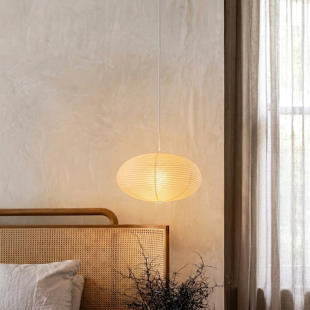 A warm, glowing oval pendant light with a ribbed paper or wicker shade hanging in a softly lit bedroom above a woven rattan headboard.