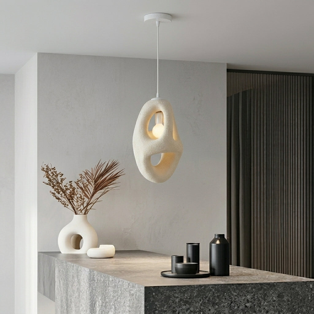 An abstract, organic-shaped pendant light in a warm cream color hangs over a modern stone countertop in a minimalist interior setting.