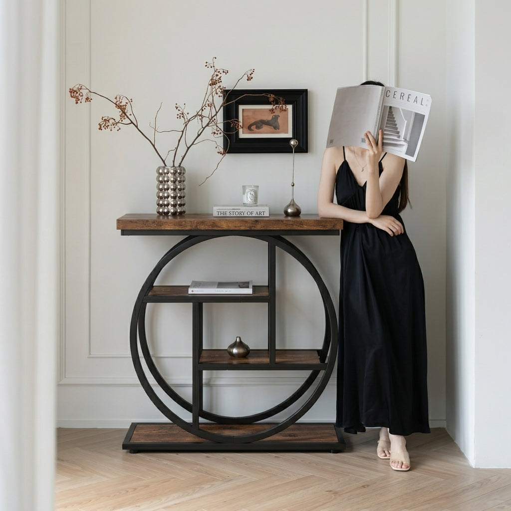 A woman in a black dress stands beside a round console table in a bright room, holding a magazine over her face. The table has a dark wood top and base with a black metal circular frame and open shelving, styled with a vase of branches, a small candle, and framed wall art.