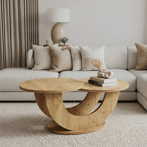 A modern round wooden coffee table with a sculptural curved base, styled in a cozy living room setting with a light beige sectional sofa and neutral decor.