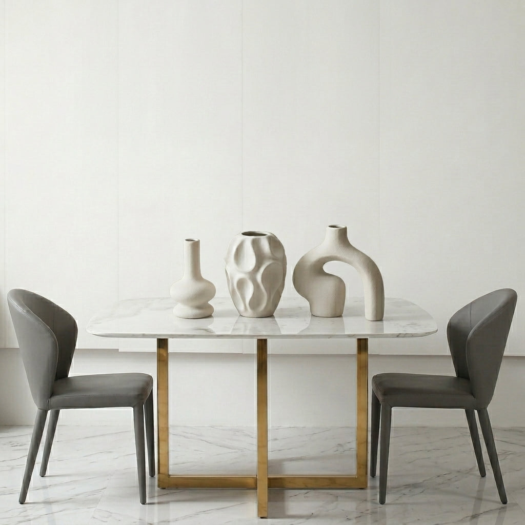 A modern dining table with a white marble top and gold metal base, flanked by two gray upholstered chairs. Three sculptural off-white ceramic vases are arranged on the tabletop against a clean white paneled wall.