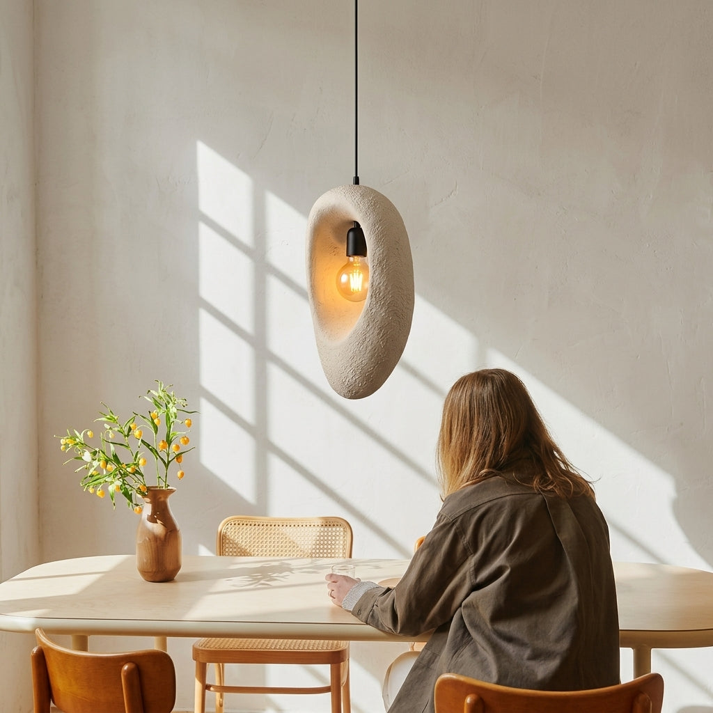 A person sits at a table in a sunlit room, viewed from behind, with a sculptural pendant light hanging above. The light fixture has a rough, organic beige stone-like shape and a visible warm bulb inside.