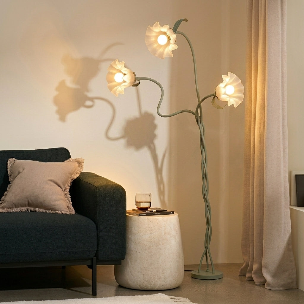 A three-light floor lamp with sculptural, flower-like white shades and a twisted sage green metal frame, styled in a cozy living room next to a dark blue sofa and a textured white side table.