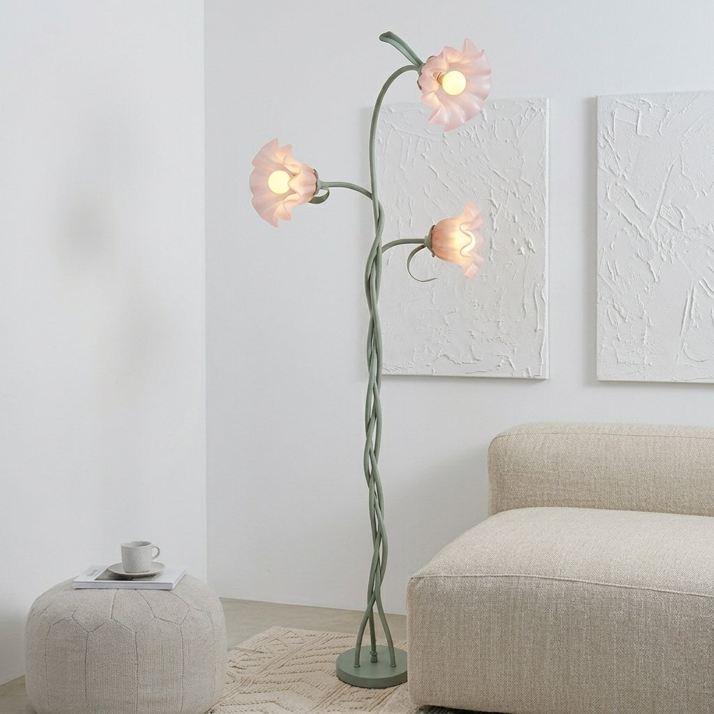 A three-light flower-shaped floor lamp with a twisted green stem and pale pink flower shades, shown lit in a modern living room setting next to a beige sofa and pouf.