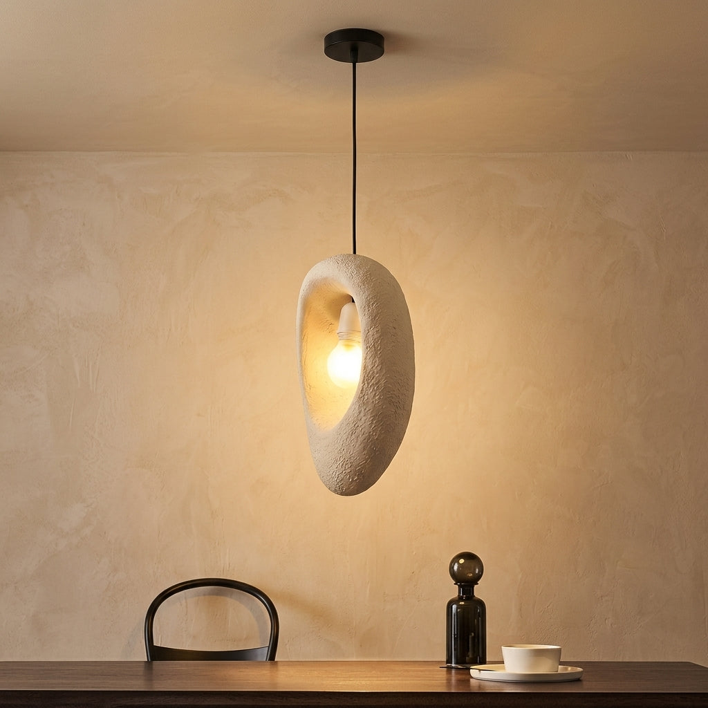 A modern pendant light with a textured, organic-shaped beige shade and a black cord, hanging in a warm, dimly lit room above a wooden table with a chair and a small bottle.