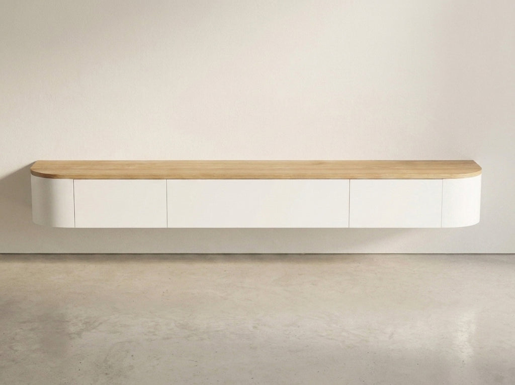 A minimalist wall-mounted floating cabinet with a light natural wood top and white curved lower body, shown against a plain off-white wall and concrete floor.
